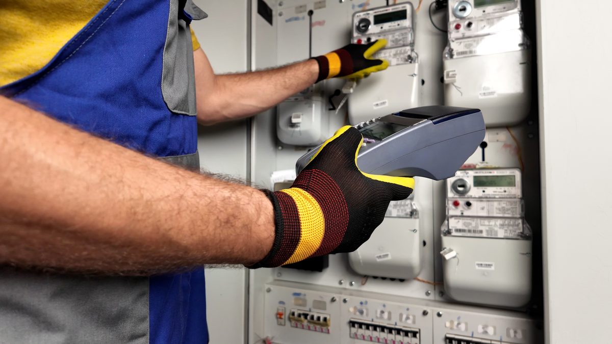 Gloved hands holding a handheld reader in front of a panel of electricity meters, with rows of circuit breakers below