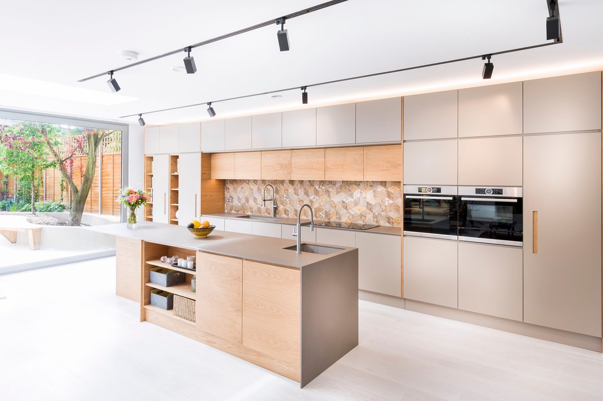 A modern kitchen with black track lighting and spotlights over a timber island, wall ovens and a garden beyond glass doors